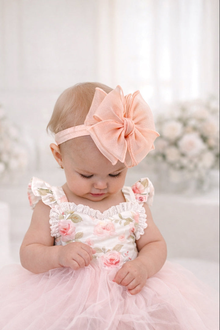 Baby in a pink floral dress with a matching headband sitting on a white surface with flowers in the background.