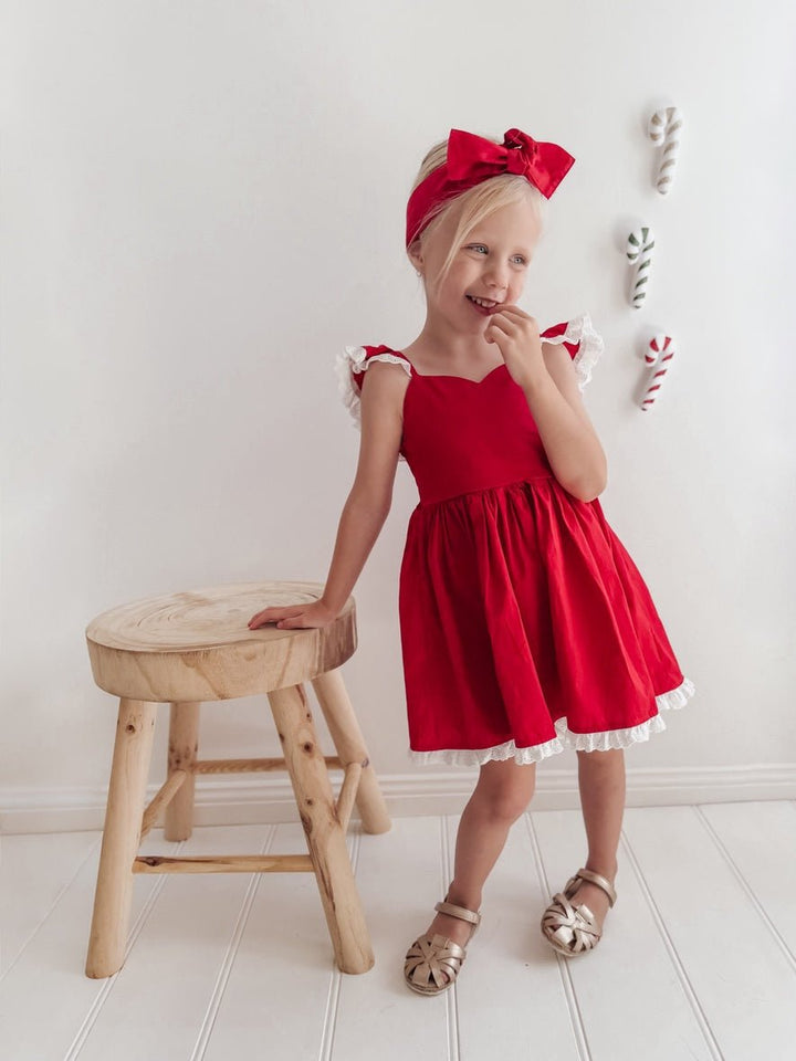 Girl wearing the Rosie red Christmas dress with white broderie trim and a matching head wrap beside a wooden stool