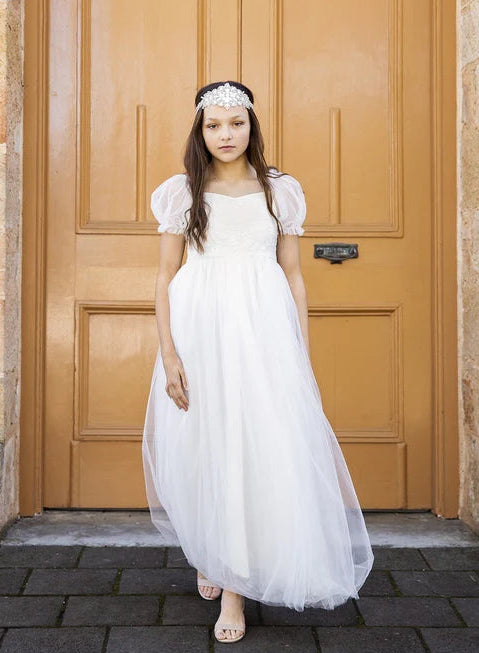 A girl wearing a white long tulle skirt dress with puffed sleeves and a soft lace bodice, standing in front of a wooden door.