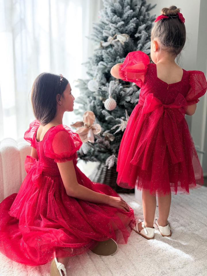 Two girls wearing Nina red sparkle Christmas dresses with puff sleeves and bow backs beside a Christmas tree