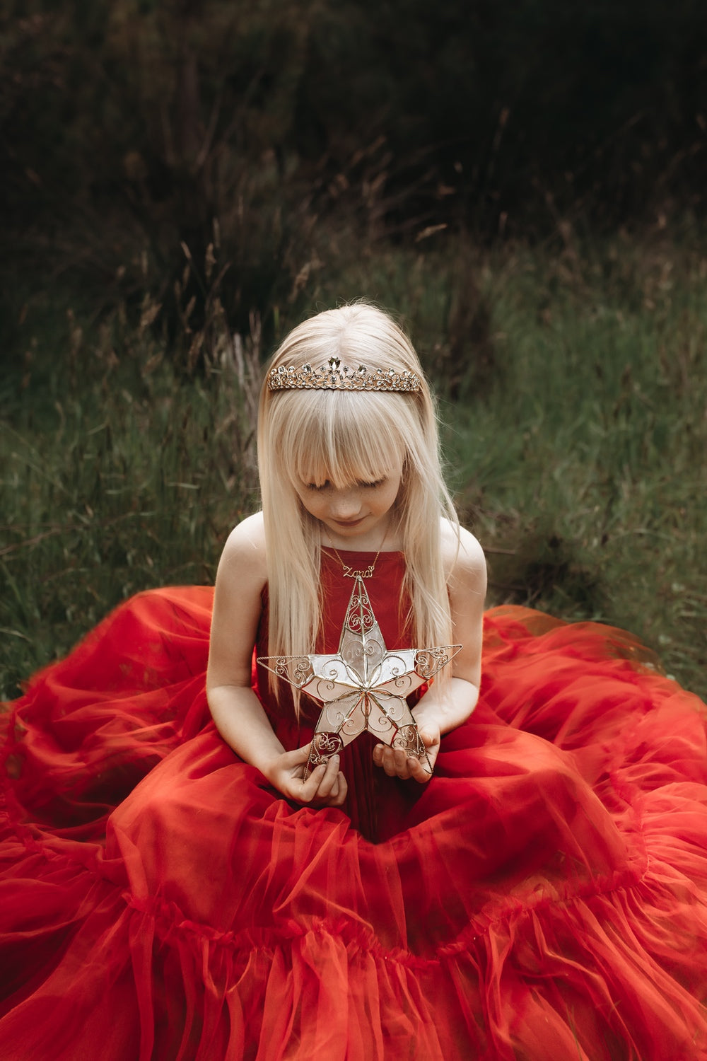 A girl in a red Christmas dress holding a gold star, she has a gold tiara in her hair.