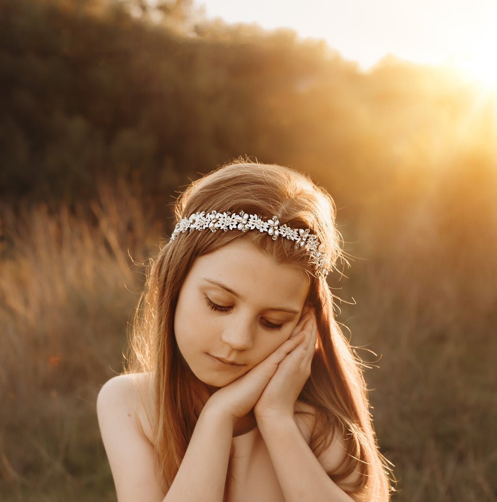 Tween girl wearing a pearl and crystal headpiece in a boho style, she is standing outdoors with the sunlight shining on her. 