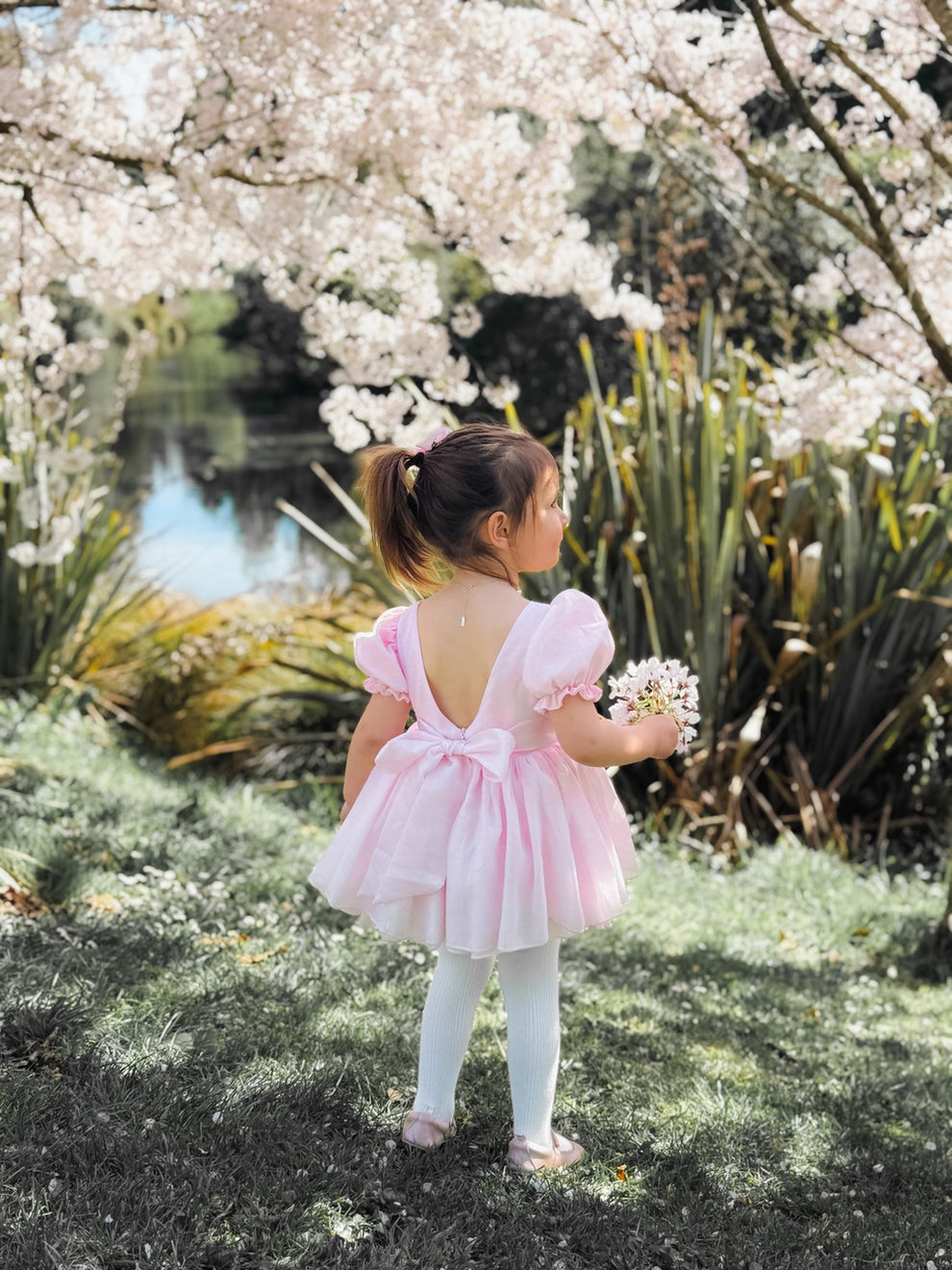 Standing sitting in Arabella baby pink dress romper, around pink flowers