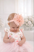 Baby in a pink floral dress with a matching headband sitting on a white surface with flowers in the background.