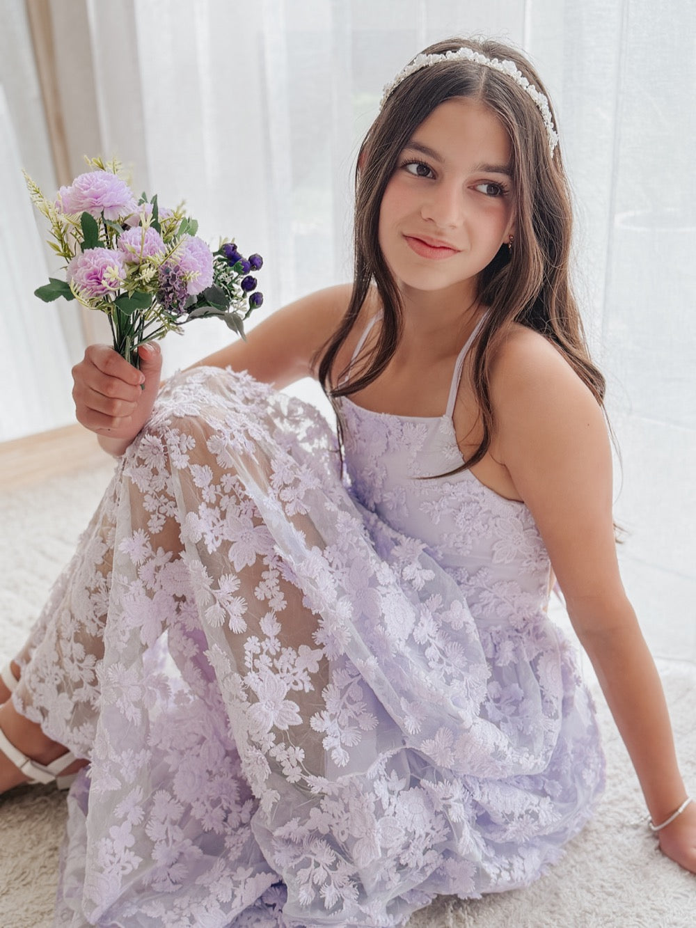 Clarissa lilac junior bridesmaid dress with 3D flowers, seated portrait with a bouquet and white headband.