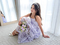 Clarissa lilac junior bridesmaid dress with 3D flowers, seated with a bouquet.