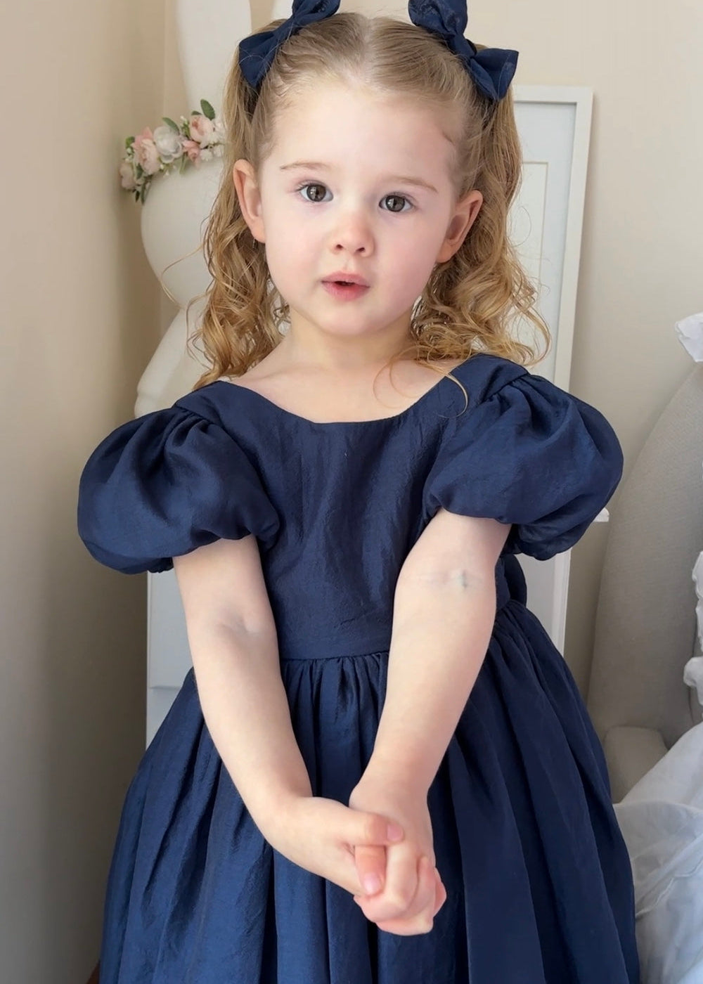Delia Girls Navy Blue Dress - Flower Girl DressesYoung girl wearing a navy blue flower girl dress with puffed sleeves in a neutral setting.
