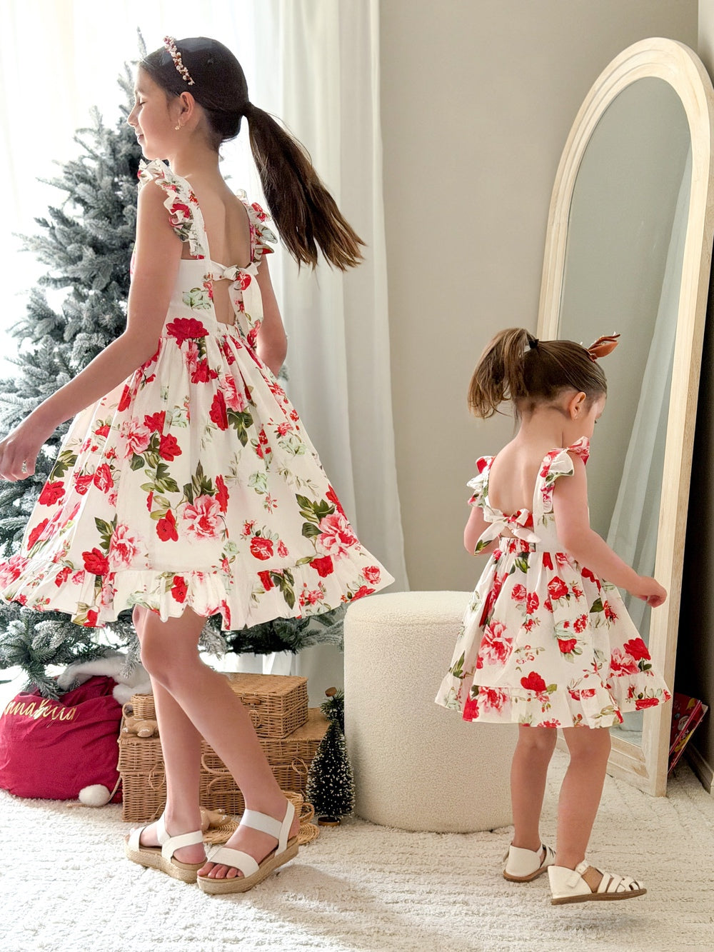 Older sister and younger sister wearing matching Faith Christmas Rose Dresses smiling by the Christmas tree
