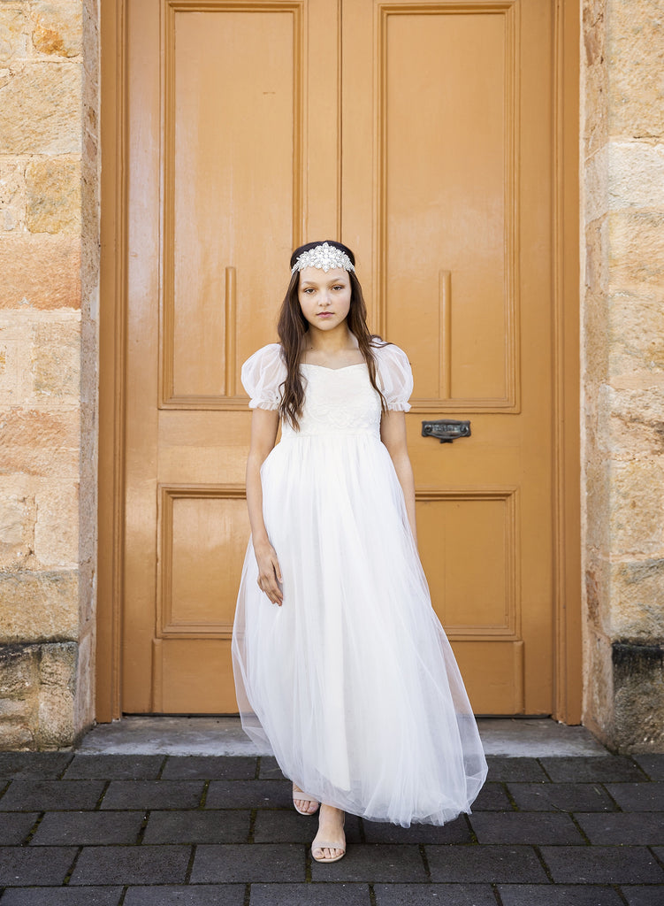 Girls White Flower Girl Dress - a girl wearing a white Communion dress standing up against a large yellow door. The dress has puffed sleeves and is a long tulle dress with lace bodice.