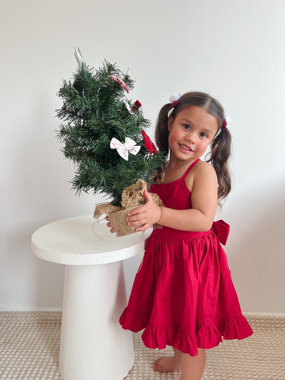 Little girl holding mini Christmas tree in red cotton Christmas dress for family holiday photos