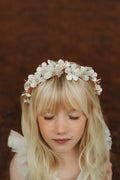 A young girl wearing a gold headband adorned with white flowers and small sparkling diamantes at their centers.