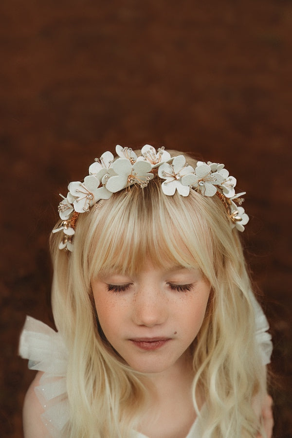 A young girl wearing a gold headband adorned with white flowers and small sparkling diamantes at their centers.