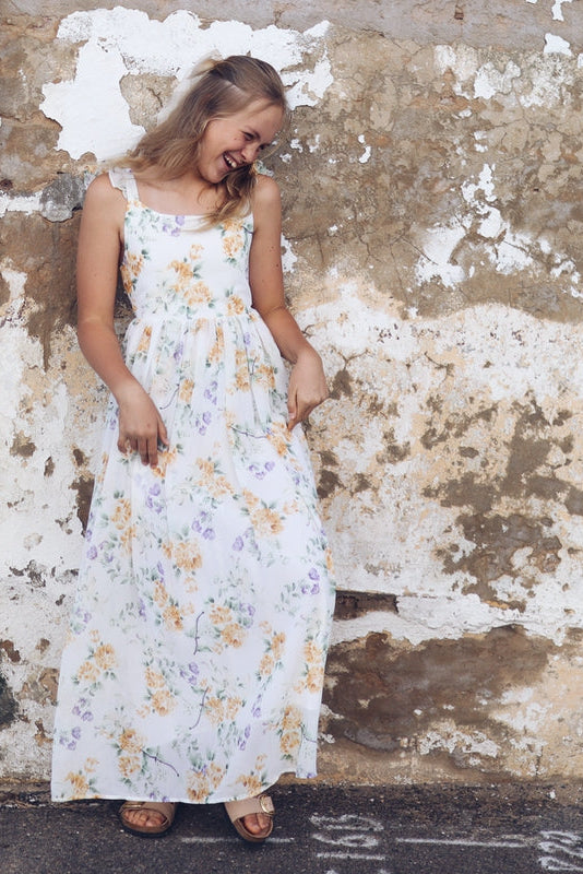 Teen girl in a floral long dress standing against a weathered wall.