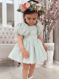 A baby girl wearing a sage green romper with puff sleeves, paired with a flower crown, standing indoors.
