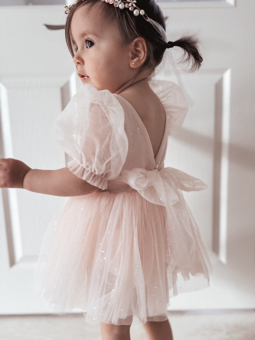 A baby girl wearing a peach-colored romper with puffed tulle sleeves and a V-back with a bow and pearl buttons.
