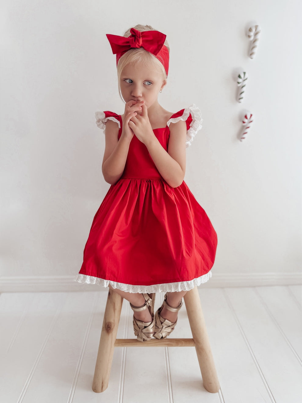 Girl seated on a stool in the Rosie red Christmas dress with white shoulder frills and a red bow head wrap