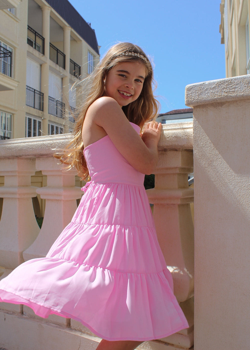 Phoebe Chiffon Girls Dress Pink - Tween Girls DressesYoung girl in a pink dress standing on a balcony with a clear blue sky.