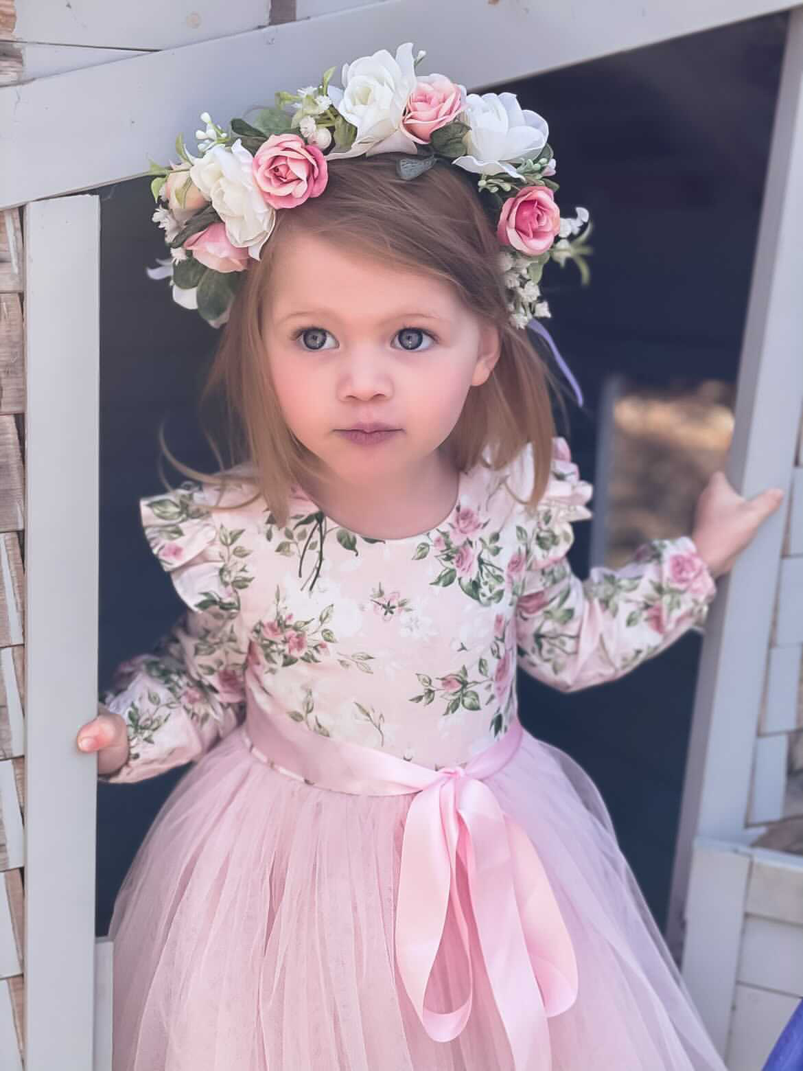 A young girl wearing a long sleeve pink floral dress with a tulle skirt and ruffle shoulders. She has a flower crown on her head.